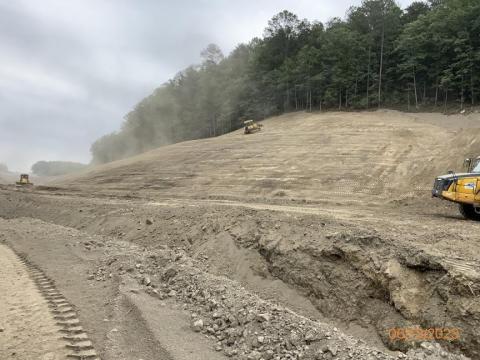 Three pieces of yellow heavy equipment stabilize a soil covered, gently sloping hillside below a treeline